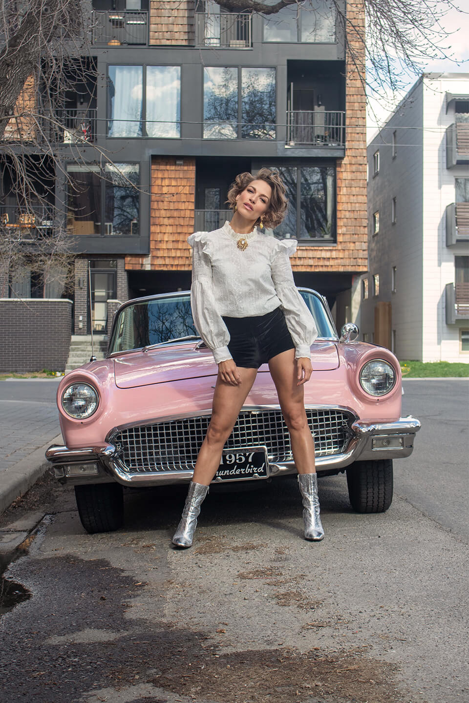 Elle McAndrews posing in front of a vintage pink 1957 Thunderbird car, wearing a stylish white blouse, black shorts, and silver boots. Urban street style meets classic car aesthetic.